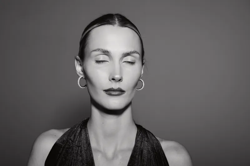 Elegant Caucasian woman with pale skin and closed eyes wears hoop earrings and dark sleeveless top in monochrome studio portrait. Concept of calm and minimalism.