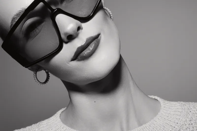 Close-up of a young white woman with pale skin wearing large sunglasses and hoop earrings, posing with a neutral expression in monochrome.