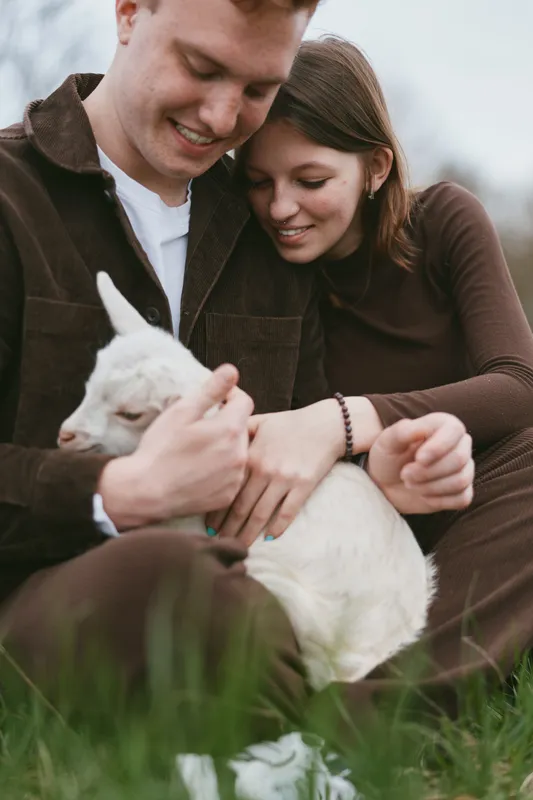 A couple tenderly cradles a white goat kid outdoors, smiling and relaxed in a sunny, grassy setting. A lifestyle moment highlighting care, companionship, and calm rural living, with gentle animal
