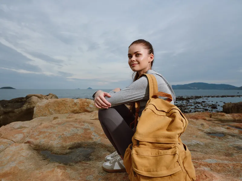 Young woman enjoying nature by the sea, wearing a casual outfit with a yellow backpack, expressing a joyful emotion against a rocky coastal background