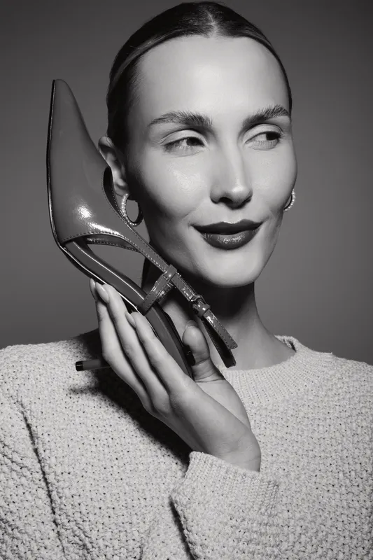 Portrait of a Caucasian woman with light skin holding a high heel shoe near her face, wearing a knitted sweater and earrings, studio lighting.