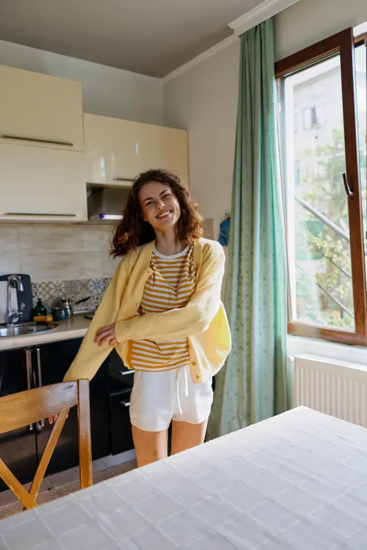 Smiling woman in a striped top and yellow cardigan standing in a bright kitchen. Casual home scene highlights everyday routine, comfort, and wellness with soft daylight and a cheerful, relaxed mood.