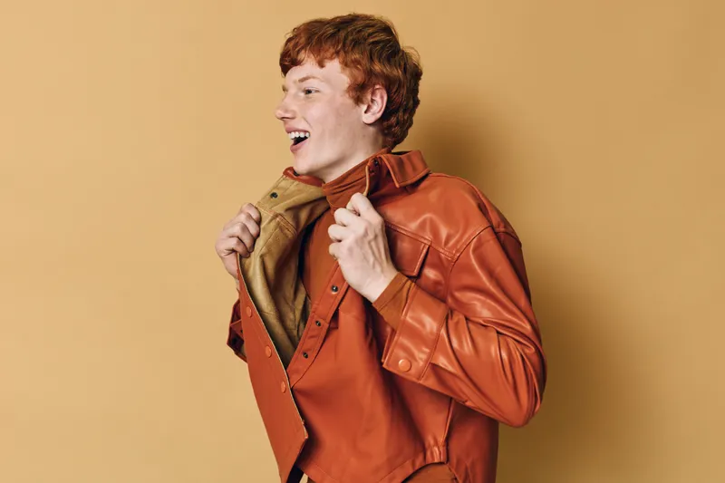 Young man with red hair wearing orange leather jacket smiles and poses with closed eyes on beige background. Casual style, happiness and confident expression with fashion mood.