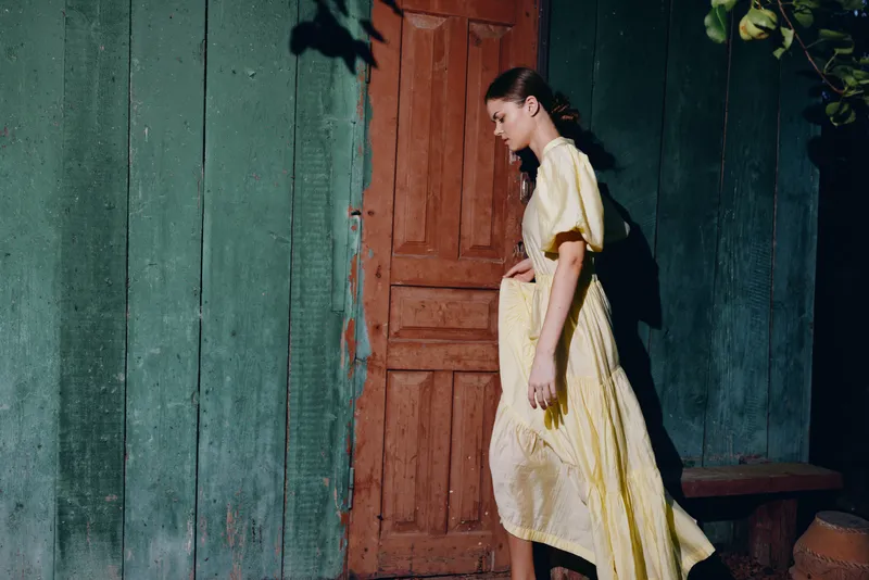 Woman in a yellow dress walks along a weathered green wall and brown door, captured in soft natural light for a moody artistic street fashion portrait.