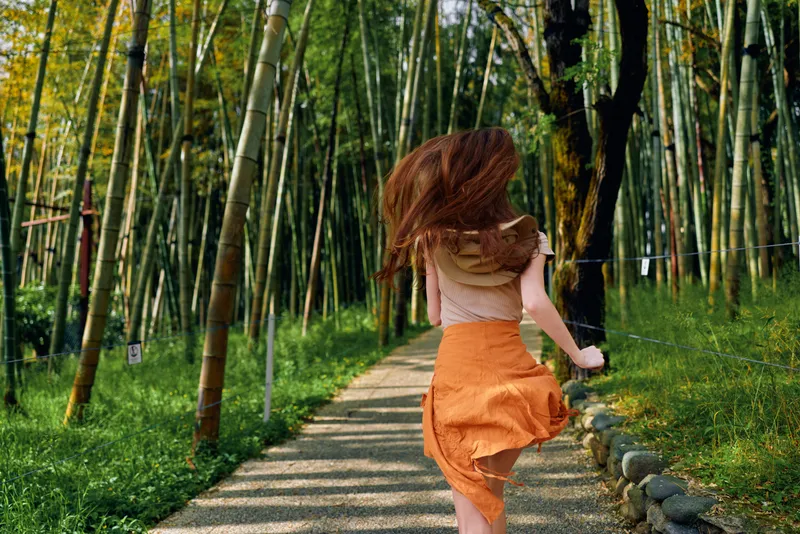 Woman running along a winding bamboo forest path in summer, orange skirt flowing, back view showing motion and carefree movement among tall green stalks on a shaded trail.