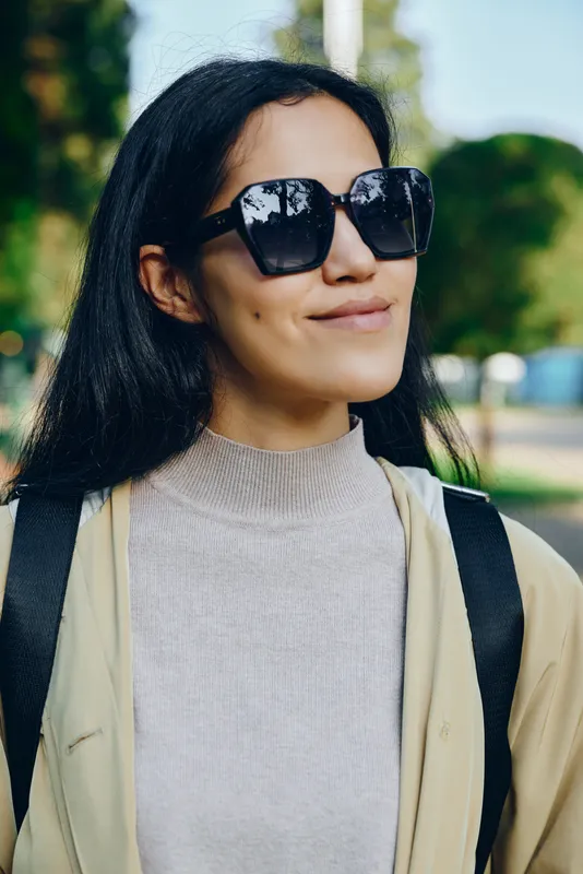 Stylish sunglasses on a woman outdoors, casual chic in bright daylight, light knit and jacket, relaxed smile, sunlit park scene, contemporary street style photography