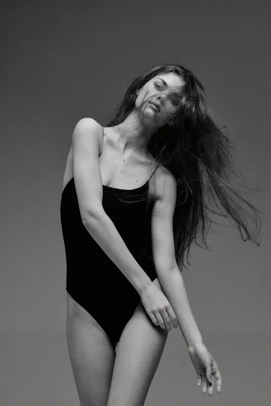 Slim woman dancing joyfully in black swimsuit with flowing hair on gray background in black and white style