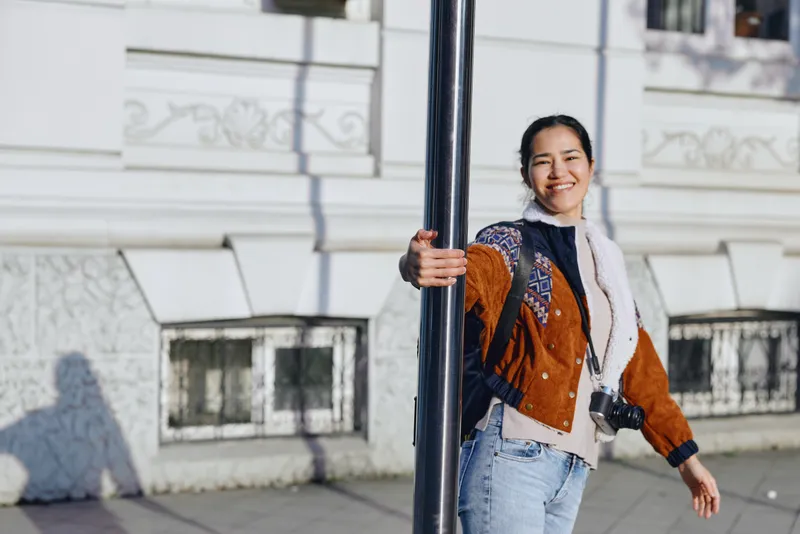 Smiling woman leaning against a street lamp in an urban setting, casual jacket and jeans, sunny day, candid street photography vibe with fashion flair and everyday movement