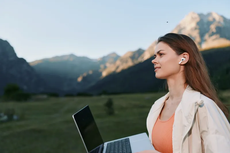 A focused female professional uses a laptop outdoors, wearing wireless earbuds. She appears attentive and confident, enjoying fresh air and natural scenery while working.