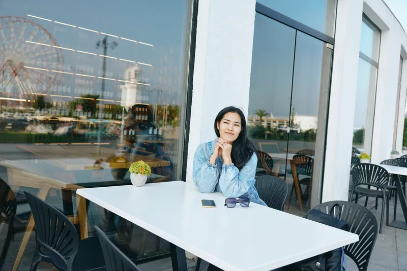 Woman sits at an outdoor cafe table by a glass storefront, daytime leisure scene, thoughtful pose, smartphone and sunglasses on the table, calm urban mood