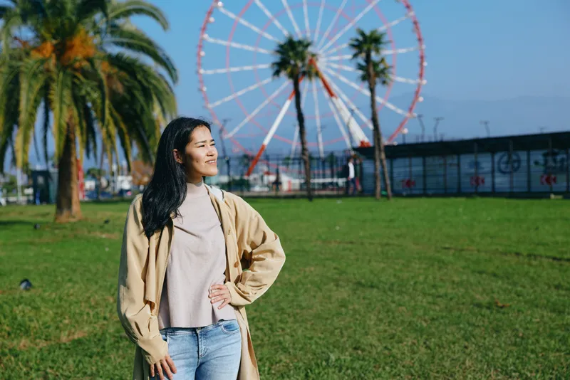 Woman wearing a casual sweater stands in a sunny park with a ferris wheel in the background, showcasing leisure, fashion, and bright outdoor photography