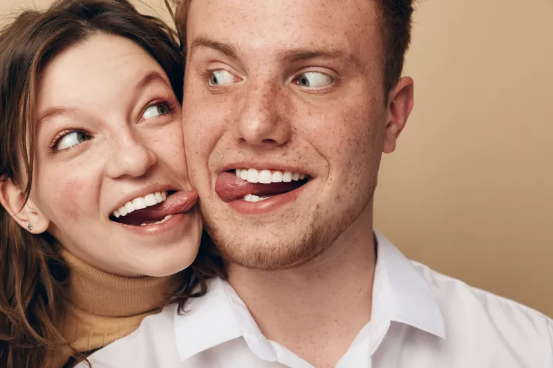 Young couple with playful expressions sticking out tongues, close together on beige background. Concept of fun, friendship, love, and joyful relationship moments between two people.