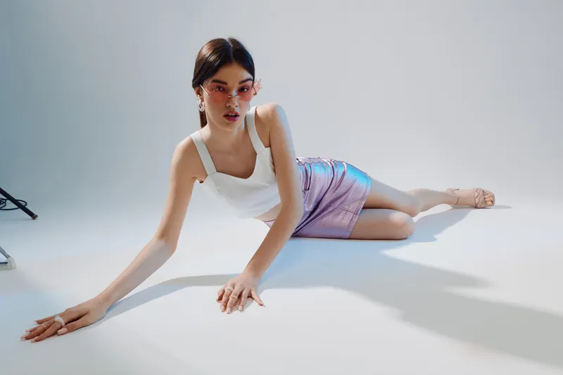 Young woman in a white top and shiny purple skirt poses on the floor in a bright studio. She wears pink sunglasses and earrings. Concept of woman beauty fashion and modern style.
