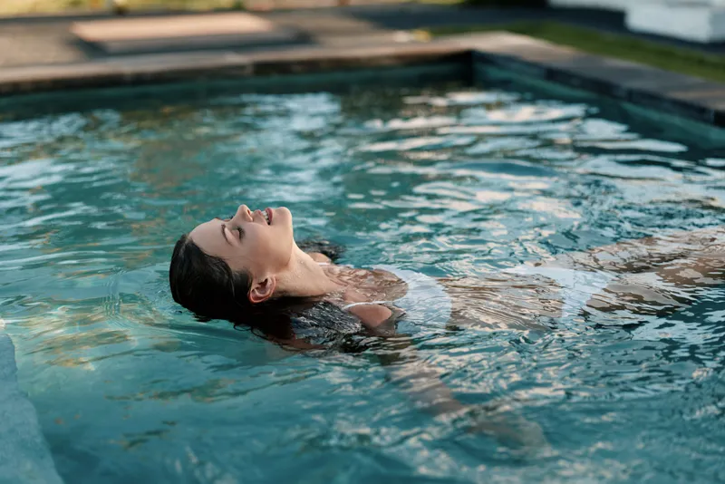 A woman floats on her back in a clear pool, eyes closed, savoring a peaceful moment. Sunlit water reflects calm, wellness, and private self care during an outdoor swim.