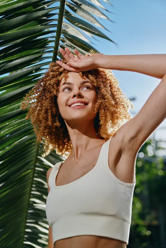smiling woman with curly hair wearing white top shielding eyes from bright sunlight near tropical palm leaves outdoors in summer