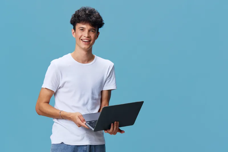 young man holding laptop smiling, casual clothing, standing against blue background, cheerful expression, technology usage, modern lifestyle portrait