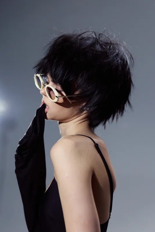 Stylish woman wearing sunglasses with messy hair in black clothing, posing thoughtfully against a minimalist gray background.