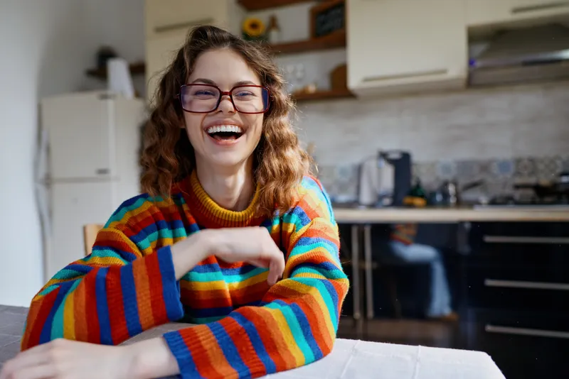 Young woman with curly hair smiles brightly at a sunlit kitchen table, wearing a colorful striped sweater, as warm daylight and a casual domestic scene convey a relaxed, joyful home moment.