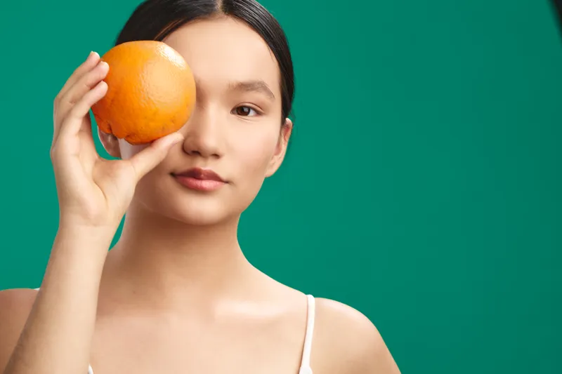 Asian woman holds orange near eye in a studio portrait with a vivid green background, creating a playful, fresh mood through simple composition, soft lighting, and a calm, minimal wardrobe.