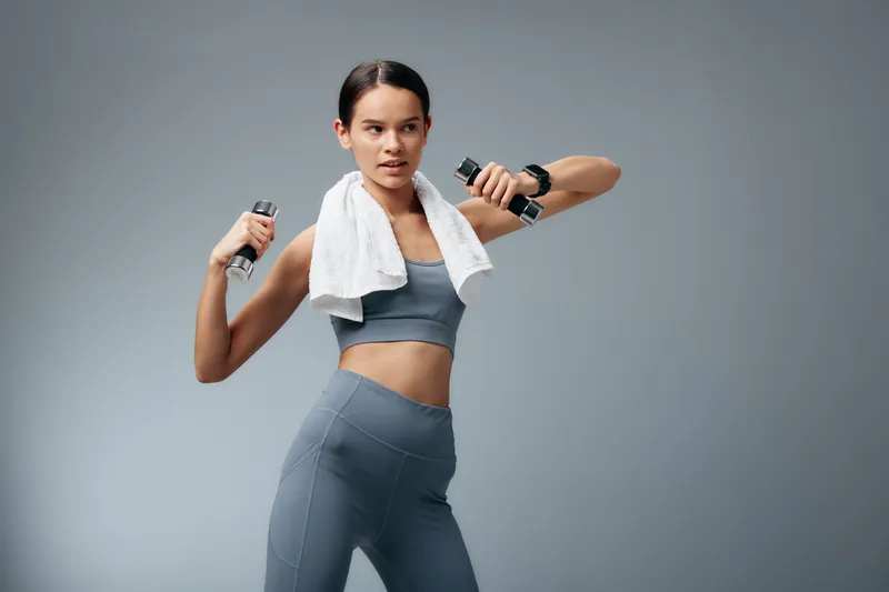 Fitness woman in athletic wear trains with dumbbells, towel draped around shoulders, mid workout in a clean studio, showcasing strength, balance and focus during a dynamic full body routine.
