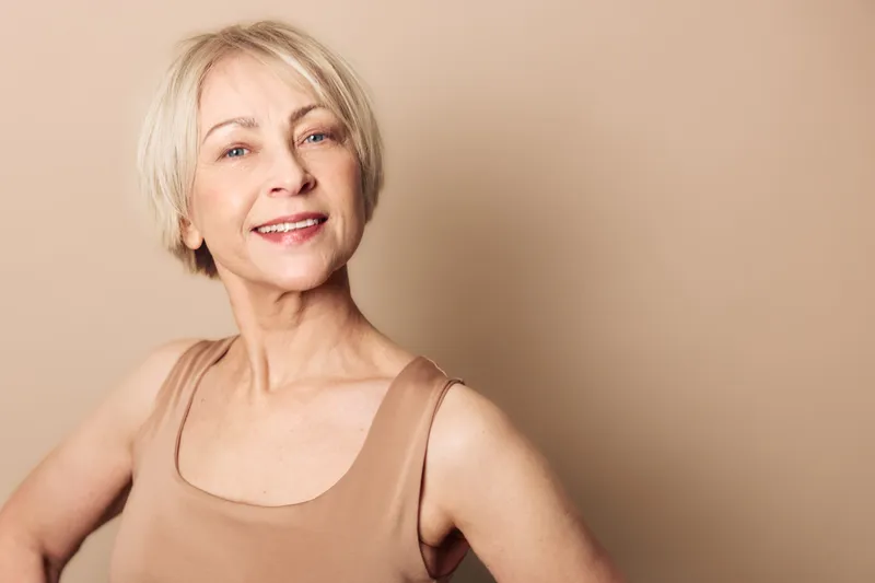 Smiling mature woman with short blonde hair wearing beige tank top poses confidently against light brown background. Concept of aging beauty, confidence, natural skin, and positive lifestyle.