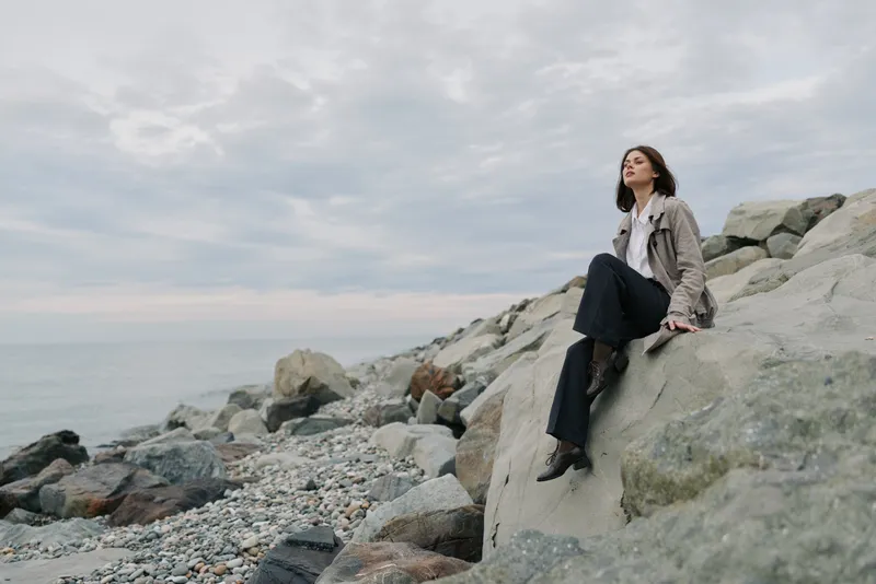 Young woman in casual outfit sitting on large rocks by the sea under cloudy sky. Concept of solitude, contemplation, and natural coastal landscape with calm mood.