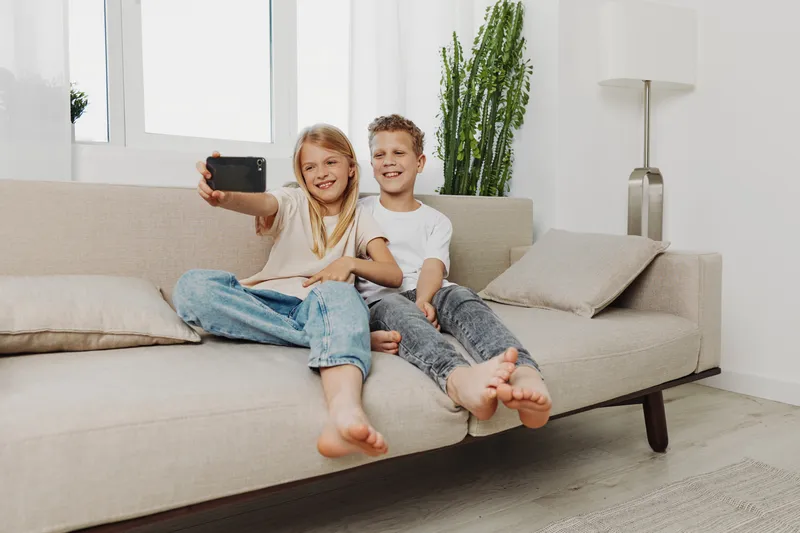 Two smiling children sitting barefoot on beige sofa in bright living room taking selfie with smartphone. Casual clothes and relaxed atmosphere create happy family moment concept.