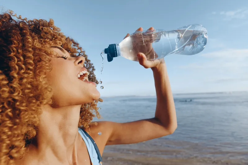 Curly woman drinking water from a plastic bottle at the beach on a sunny day. Healthy lifestyle concept with clear blue sky and ocean in the background.