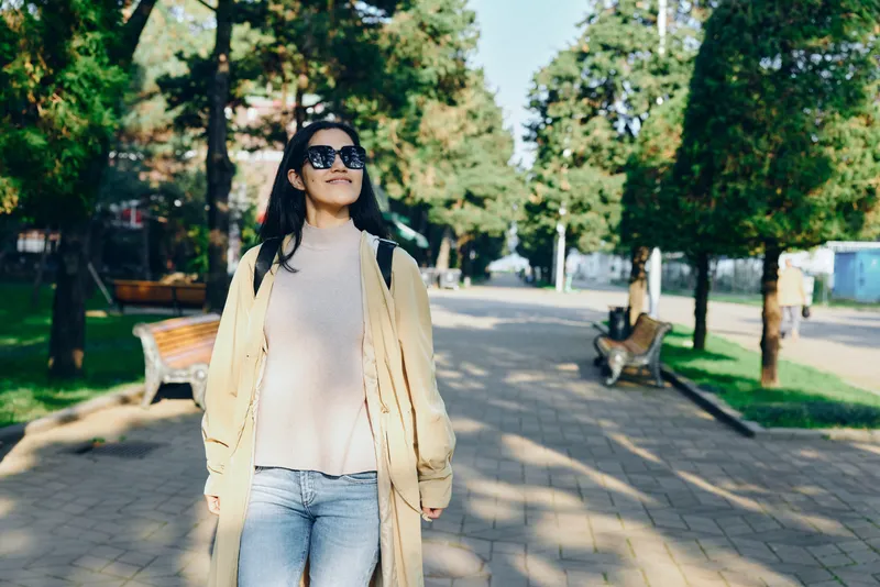 Woman in a light coat and jeans walks through a sunlit park, wearing sunglasses and a relaxed smile, enjoying a casual outdoor moment and a carefree urban stroll.