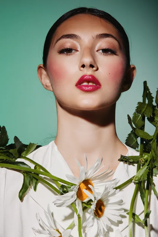 Portrait of a stylish young woman with bright red lipstick, perfect makeup, and blush. She holds daisies in a fashion beauty creative concept on a light teal background.