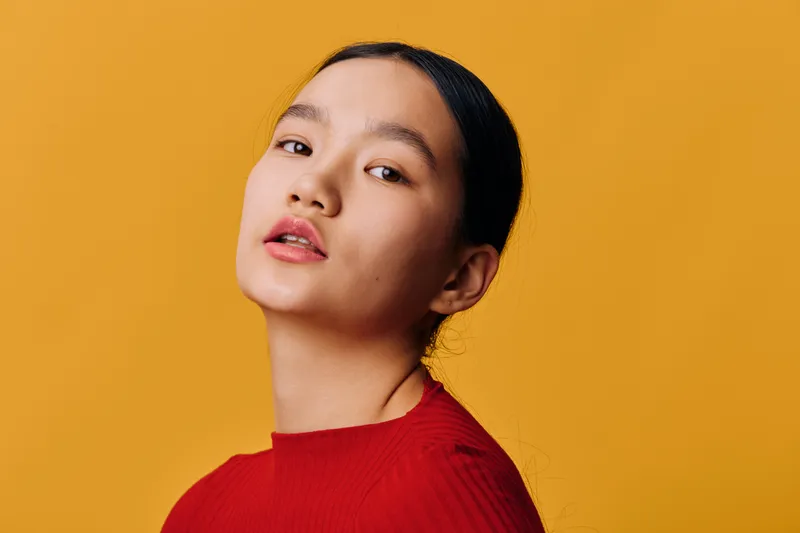 Portrait of young woman with natural makeup and dark hair tied back wearing red top against yellow background for beauty, fashion, and lifestyle concept and studio photography.