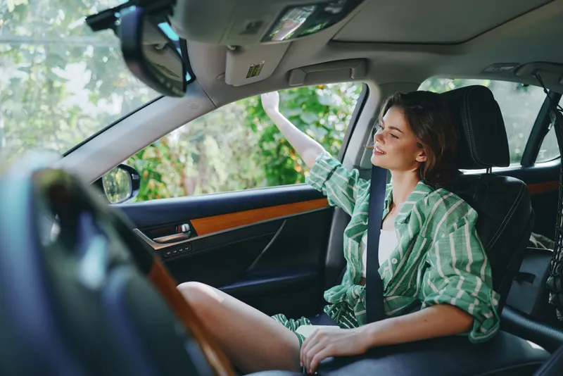 Young woman sits in the driver's seat, wearing a green striped shirt with a belt, relaxed pose inside a car during daytime, focus on comfort and style.