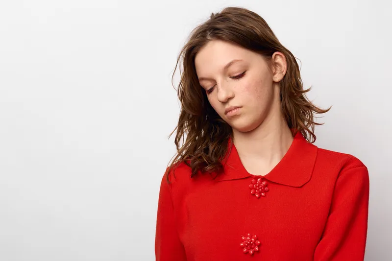 Sad young woman in red sweater looks down with closed eyes on white background. Concept of loneliness, sadness, and emotional struggle in casual clothing portrait.