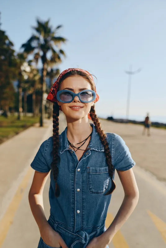 Lifestyle woman in modern boho-western denim street style wearing oversized blue sunglasses and a red bandana, captured with warm film color tones outdoors.
