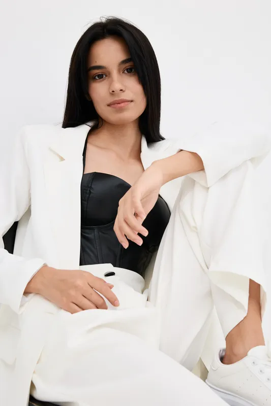 Young woman wearing white suit and black corset sitting relaxed against white background, showcasing modern fashion style with confident and calm expression.
