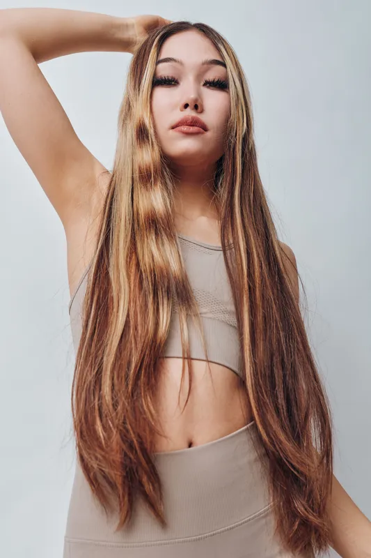 Young woman with long hair wearing beige top and leggings posing indoors with confident expression and hand in hair on neutral background.