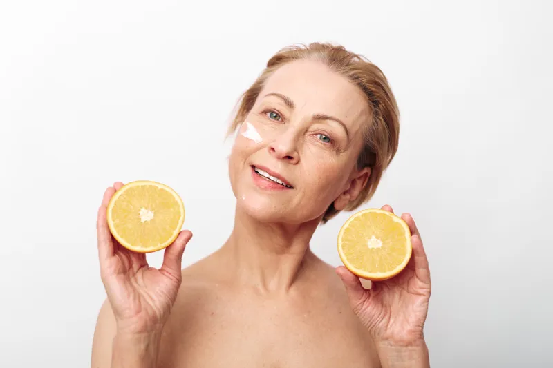 Smiling mature woman with natural skin holds two halves of fresh orange fruit near face. Wellness and healthy lifestyle concept with citrus fruits and skincare on white background.