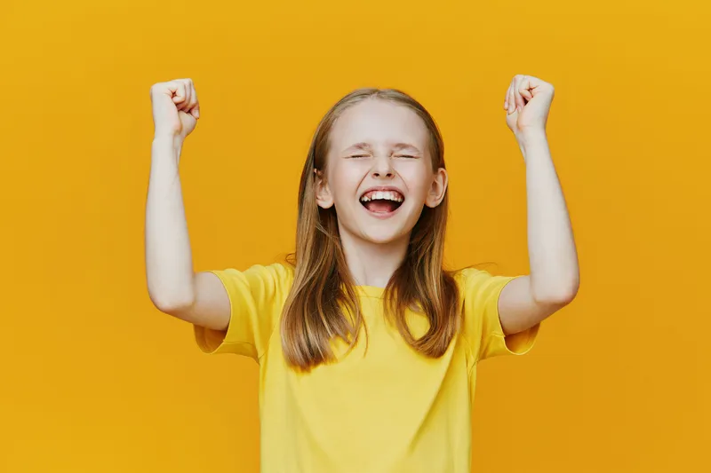 Happy girl celebrating success with joyful expression, wearing a yellow shirt against a bright yellow background, representing positivity and achievement