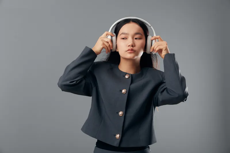 Young woman adjusting headphones with both hands wearing dark jacket studio portrait isolated on gray background. Female model posing with serious expression looking aside for music or sound concept.