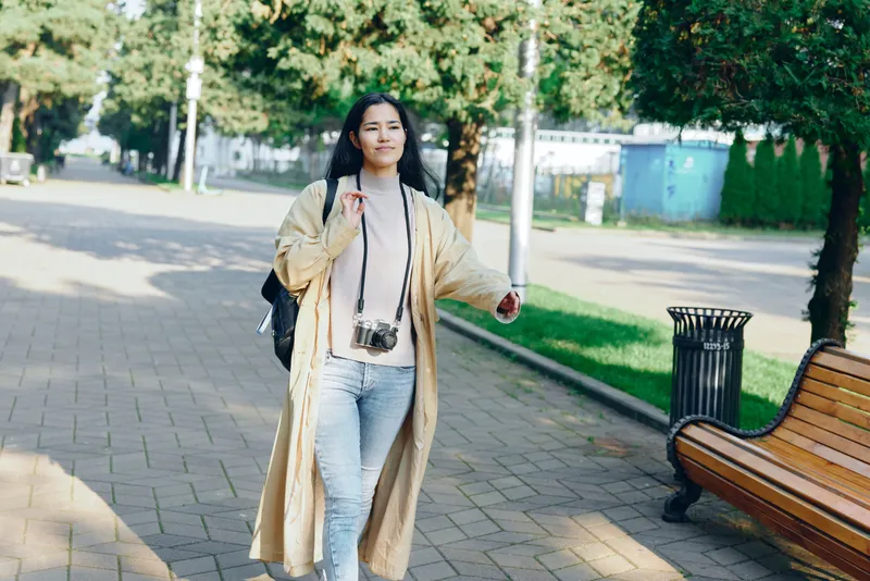 Woman strolling in a sunny park wearing a long beige coat and jeans, with a camera hanging from her neck and a backpack on her shoulder, showcasing casual outdoor fashion and relaxed urban style.