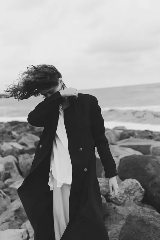 Black and white scene of a figure on a windswept beach, long coat and flowing dress in motion, moody style amid rocks and open sky at sea