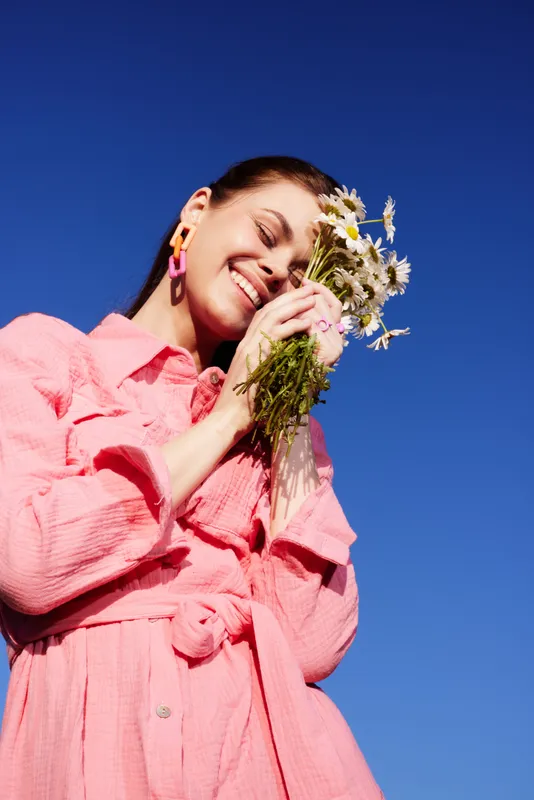Cheerful young woman holding a bouquet of daisies against a clear blue sky, wearing a bright pink dress, radiating joy and warmth during spring season