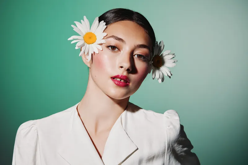 Young woman in white blouse with glossy red lips and two white daisies in hair poses confidently against green backdrop in creative fashion beauty portrait styling.