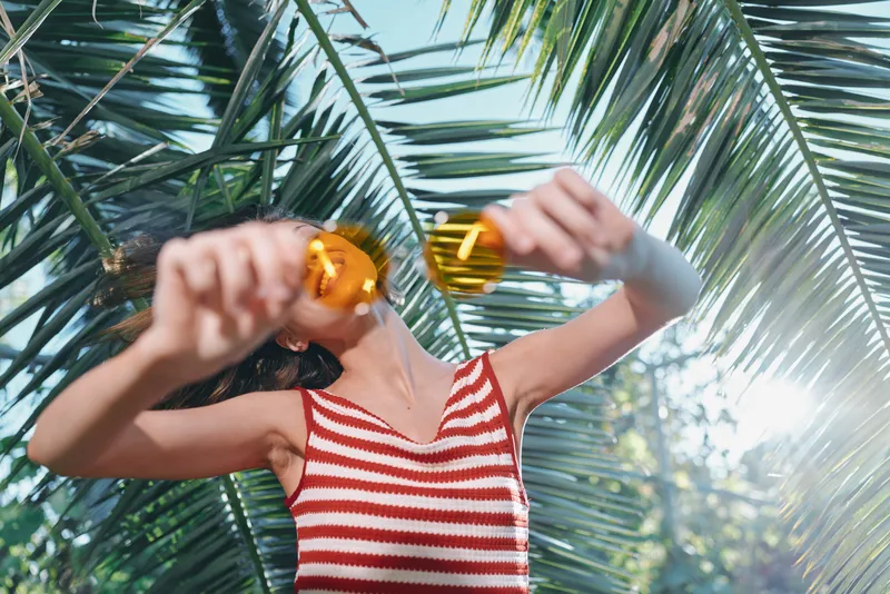 A casual figure wearing a red and white striped tank top smiles while adjusting orange sunglasses in a sunlit tropical setting, with palm fronds and bright light.