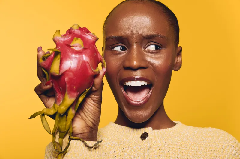 Happy black African woman with dark skin holds exotic dragon fruit close to her face, smiling and looking sideways. Bright yellow background and casual beige sweater create warm mood.