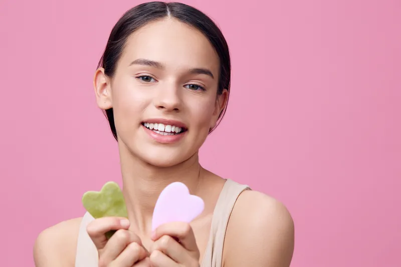 Young smiling woman with natural beauty holding heart shaped skincare products on a pink background, representing self care and wellness