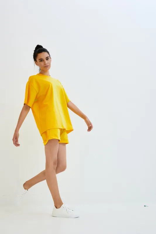 Young woman in yellow outfit walking confidently against white background, wearing casual shorts and T-shirt with white sneakers, minimalistic studio setting.