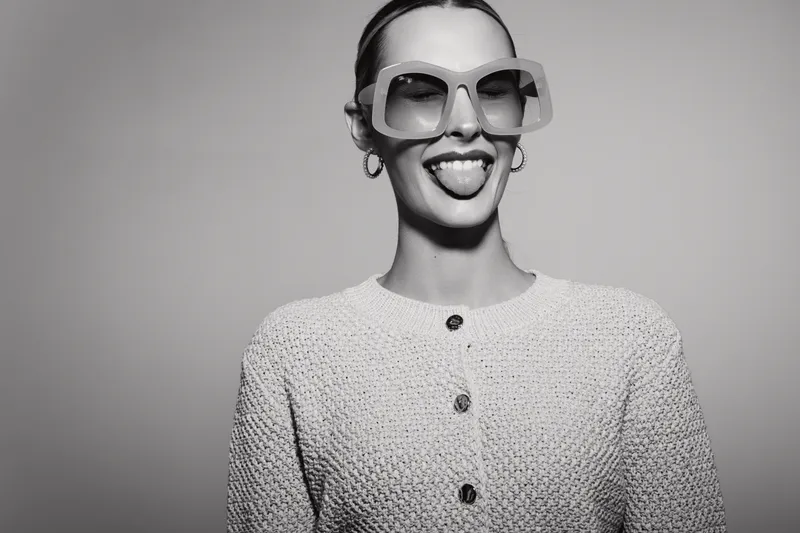 Young white woman with light skin wearing large sunglasses and earrings sticks out tongue in studio portrait.