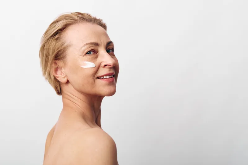 Mature woman with short blonde hair applies white face cream on cheek, smiling gently. Clean skin care concept with natural beauty and healthy lifestyle on plain white background.