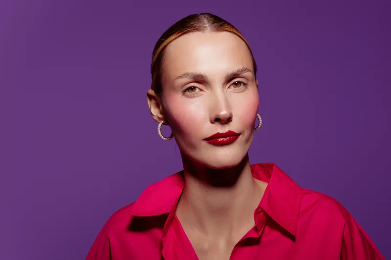 Portrait of confident woman with slicked hair wearing red shirt and hoop earrings on purple background. Studio lighting highlights facial features and makeup details.